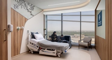 Two people are sitting and interacting beside a hospital bed in a modern hospital room with large windows