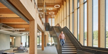 Interior shot of a building with mass timber detailing, a staircase, floor-to-ceiling windows bringing in natural light, and halo light fixtures suspended from the ceiling