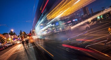 This image depicts a bustling city street at twilight with long exposure photography capturing motion blur of vehicles. Streaks of light from car headlights and tail lights create vibrant lines across the frame. Buildings and illuminated signs line the background, contributing to the urban atmosphere. The scene conveys a sense of dynamic movement and urban activity.