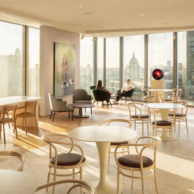 Office interior at 22 Bishopsgate, London. Tables and chairs in communal area, with 2 women sat in background in front of the London skyline showing out of the window.