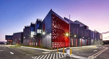 The image depicts a modern data center, NextDC's Merlot 3, featuring a striking red and gray facade, illuminated by external lights at dusk, with a clear and empty parking area in the foreground.