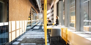 Students sitting in a light-filled hallway at a university, with yellow detailing and floor-to-ceilin windows