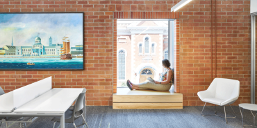 A woman sitting in a window seat inside the city of Kingston's Frontenac Central Branch Public Library. The wall is exposed brick with a work of art blue in hue showing a ship sailing in the ocean. There is a white table to the left of the window and a white chair to the right of the window.