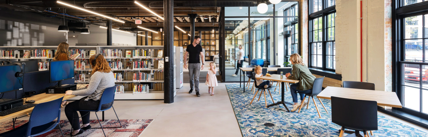 City Omaha Public Library Downtown Branch Living Room with person using computer, father walking with child, and women sitting at a table with a child in a modern library with natural lighting