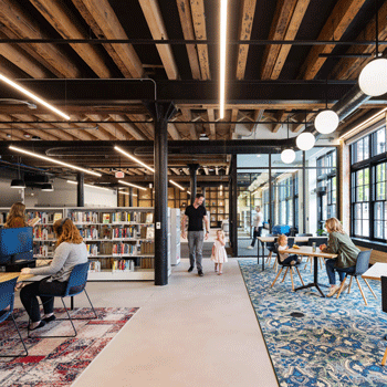 Image of the City Omaha Downtown Public Library Branch with a variety of people using the library space. One person is looking at books, another is using the computer, a father and daughter walk through the space, and a woman is sitting at a table with a child. The library floors are carpeted and the space is open and filled with daylight. 