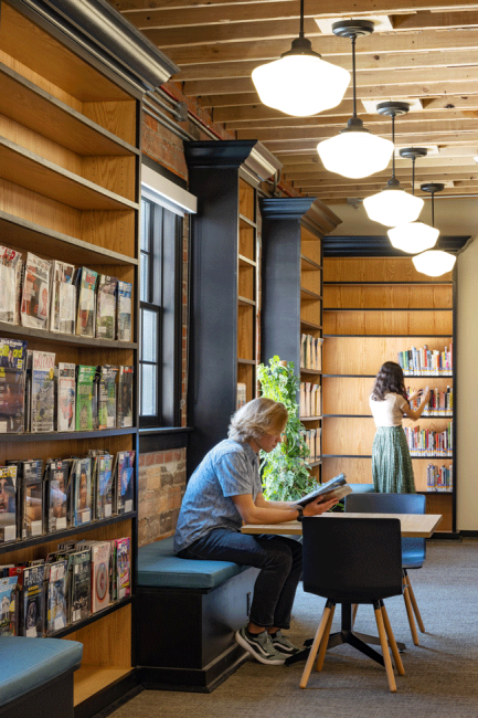 A woman reads a book at a table while another women looks through bookshelves at the Omaha Public Library Downtown Branch reading room. 