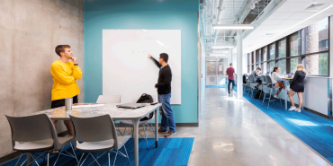 A photo of a corridor at the College Charleston Rita Hollings Science Center. On the left, two students are seen collaborating by the white board and on the right-hand side, students sit independently in bar-like seating with views to the outdoors.