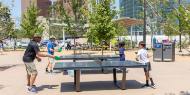 Four people playing ping pong at the Gene Leahy Mall with trees and city skyline of Omaha, Nebraska in the background.